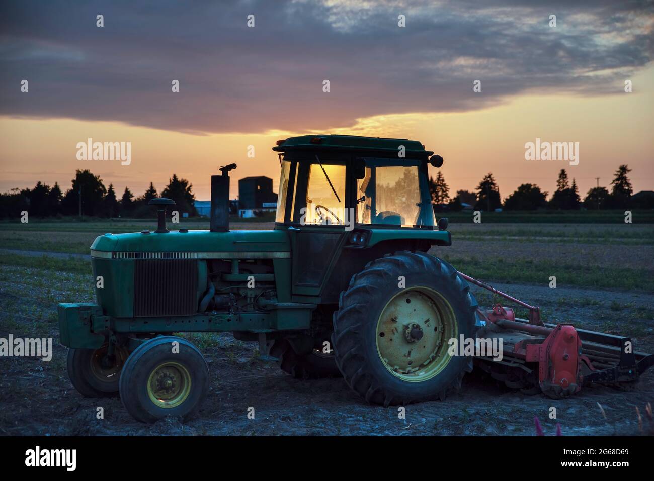 A green tractor with a hitch, illuminated by the rays of the setting sun, stands in a field on a warm summer evening. Green trees and huge sky with be Stock Photo