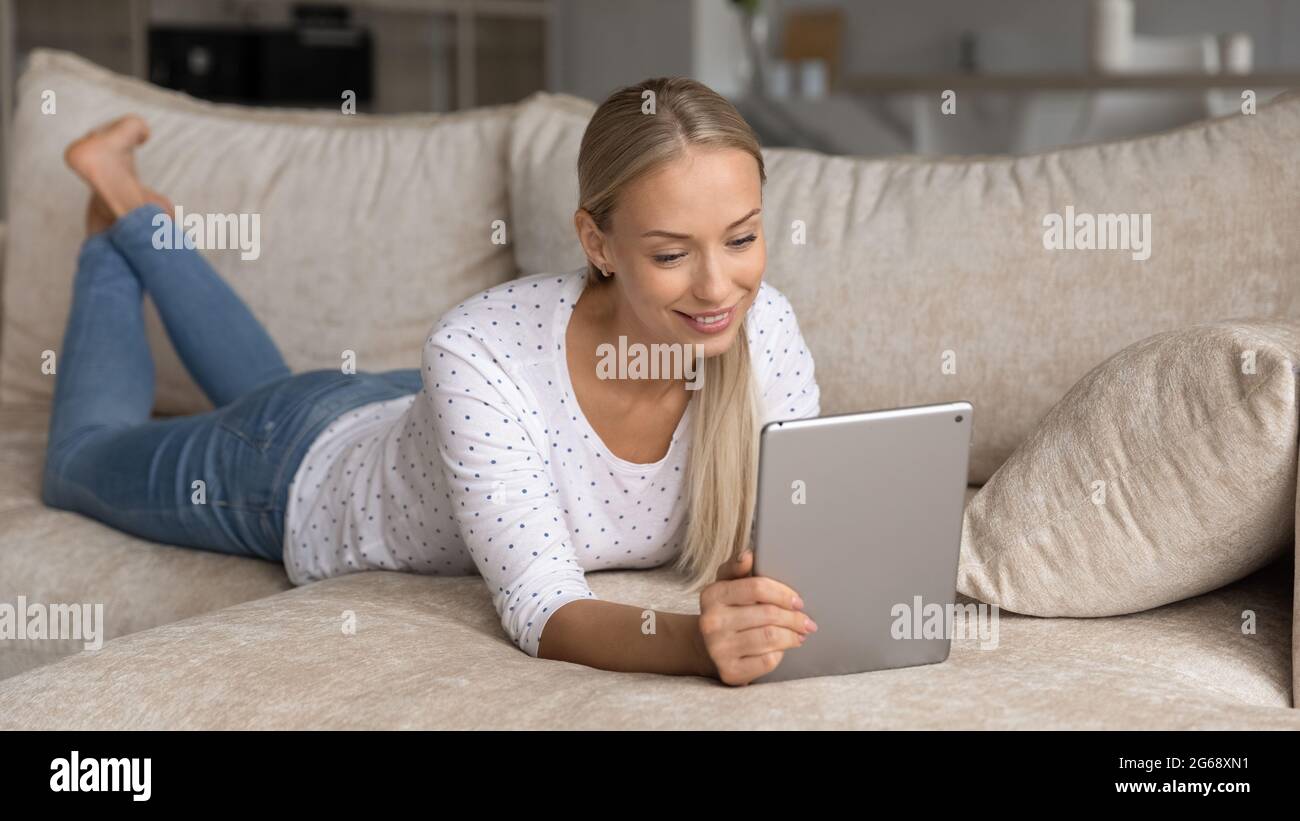 Smiling young woman using tablet, relaxing lying on cozy couch Stock Photo