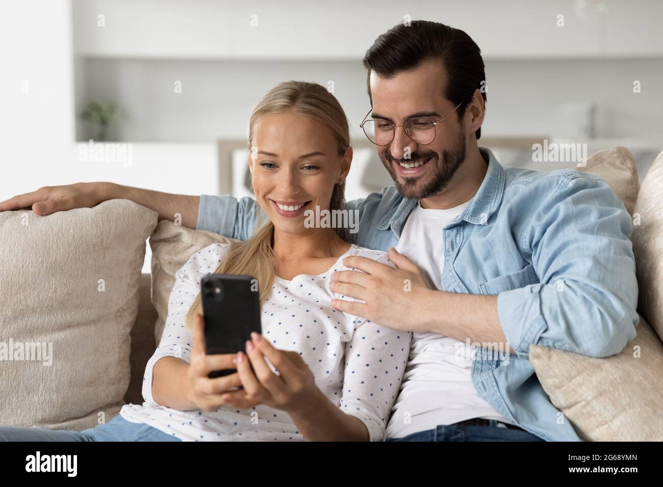 Smiling young couple hugging using smartphone sitting on couch together Stock Photo