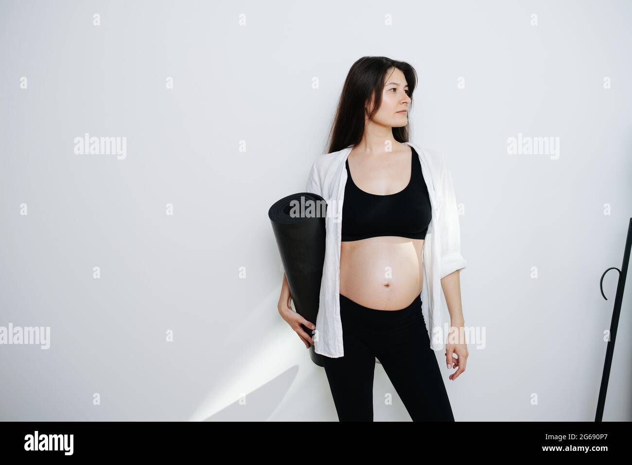 Portait of a pregnant woman holding yoga mat in hand. She's wearing black top and pants. Looking to the side. Stock Photo