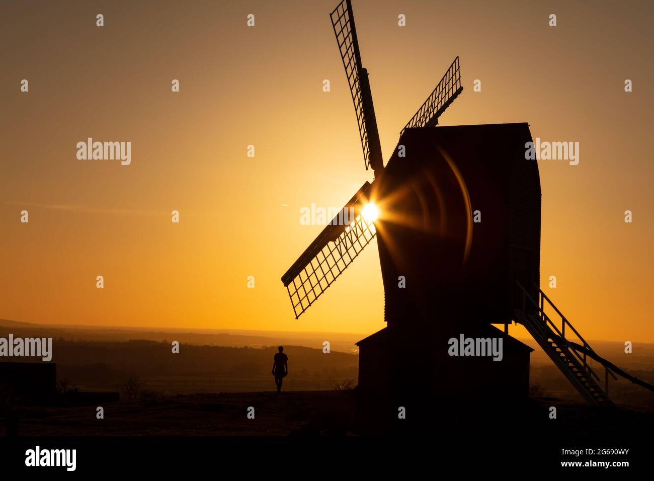 Sun setting on the horizon behind a traditional windmill at sunset with the sihouette of a man walking Stock Photo