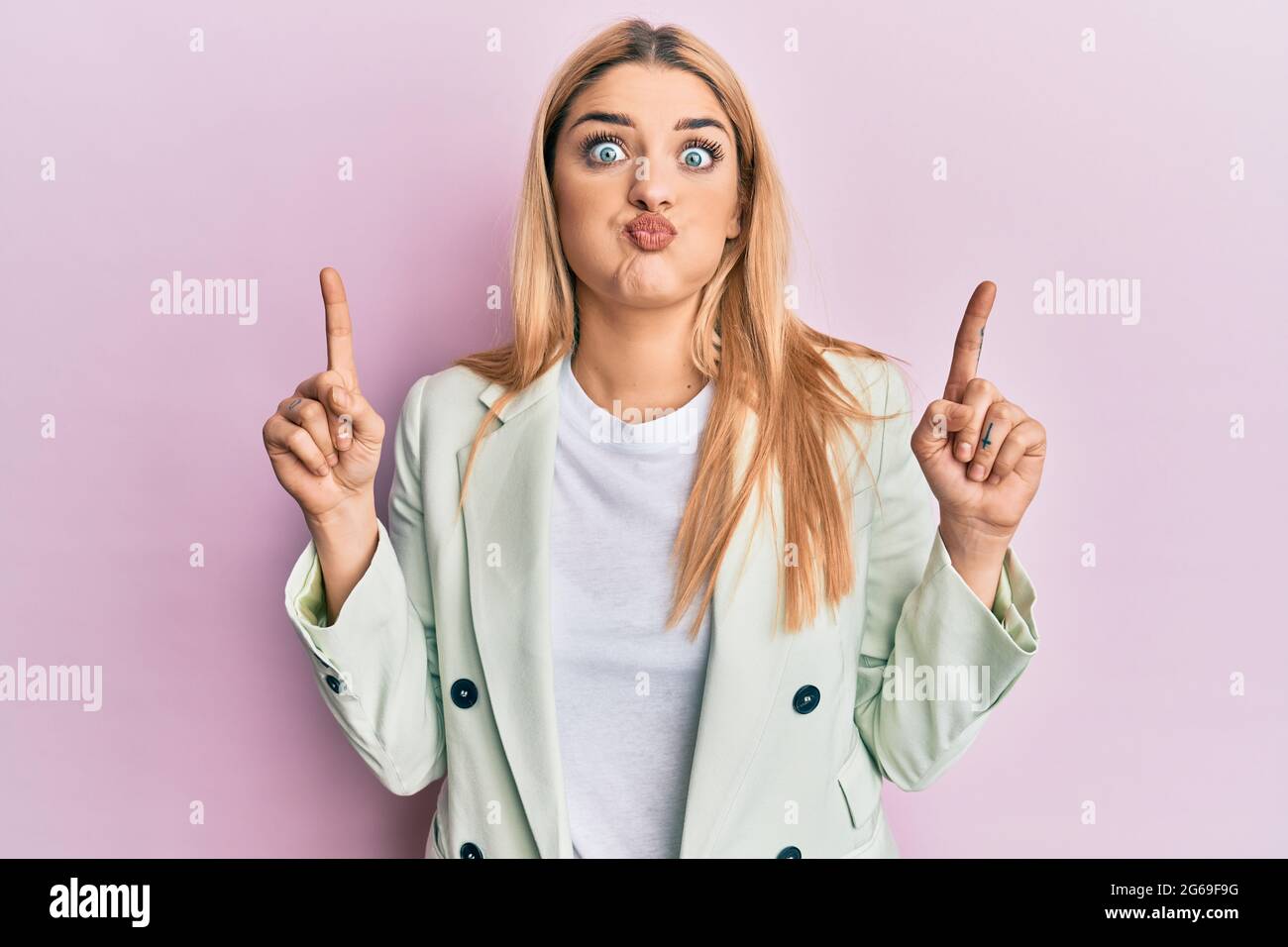 Young caucasian woman wearing business clothes pointing up with fingers puffing cheeks with funny face. mouth inflated with air, catching air. Stock Photo