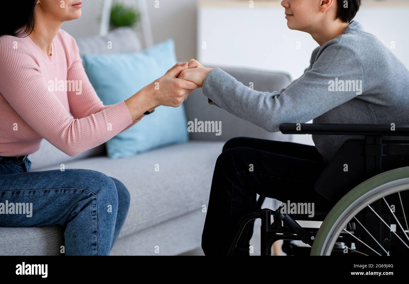 Handicapped teenage boy in wheelchair receiving support from his mother or caregiver, holding her hands at home, closeup Stock Photo