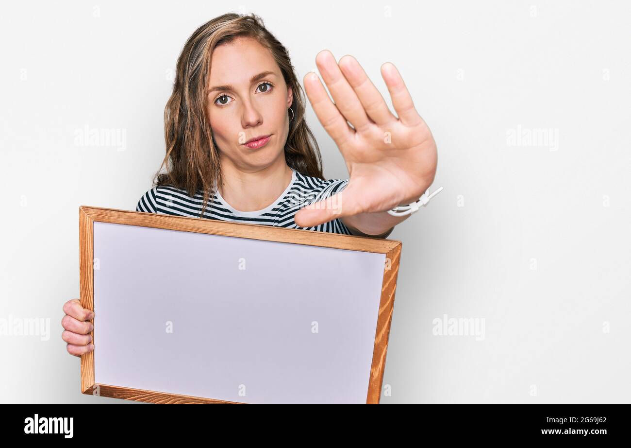 Young blonde woman holding empty white board with open hand doing stop sign with serious and confident expression, defense gesture Stock Photo