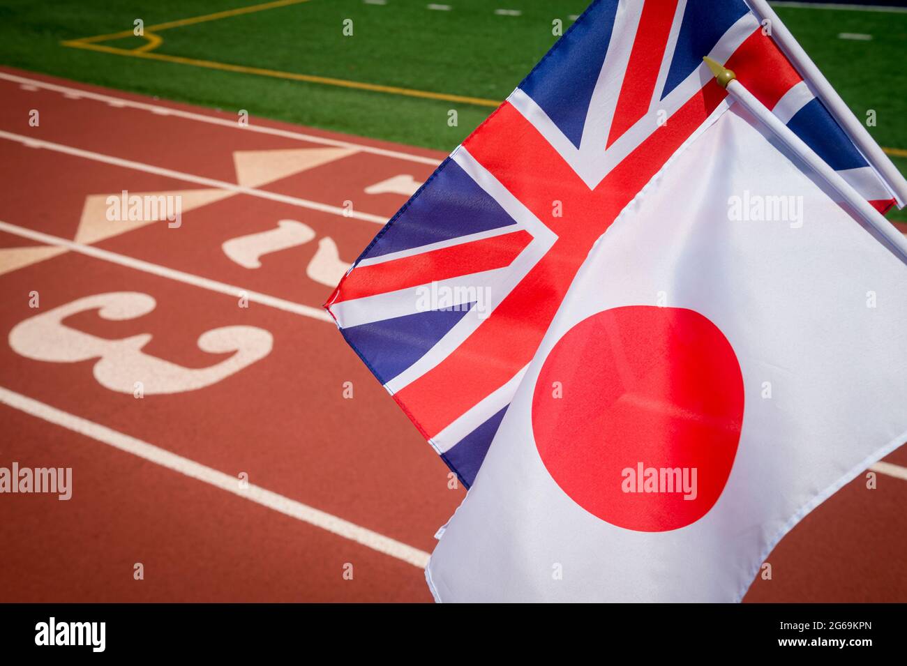 Bright sunny view of UK Union Jack and Japanese flags flying together in front of the numbered lanes at the starting line of a red running track Stock Photo