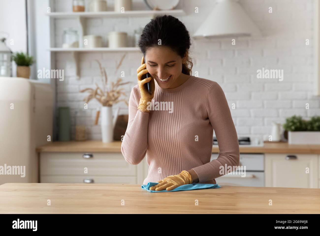 Happy young housewife chatting on cellphone while cleaning kitchen. Stock Photo