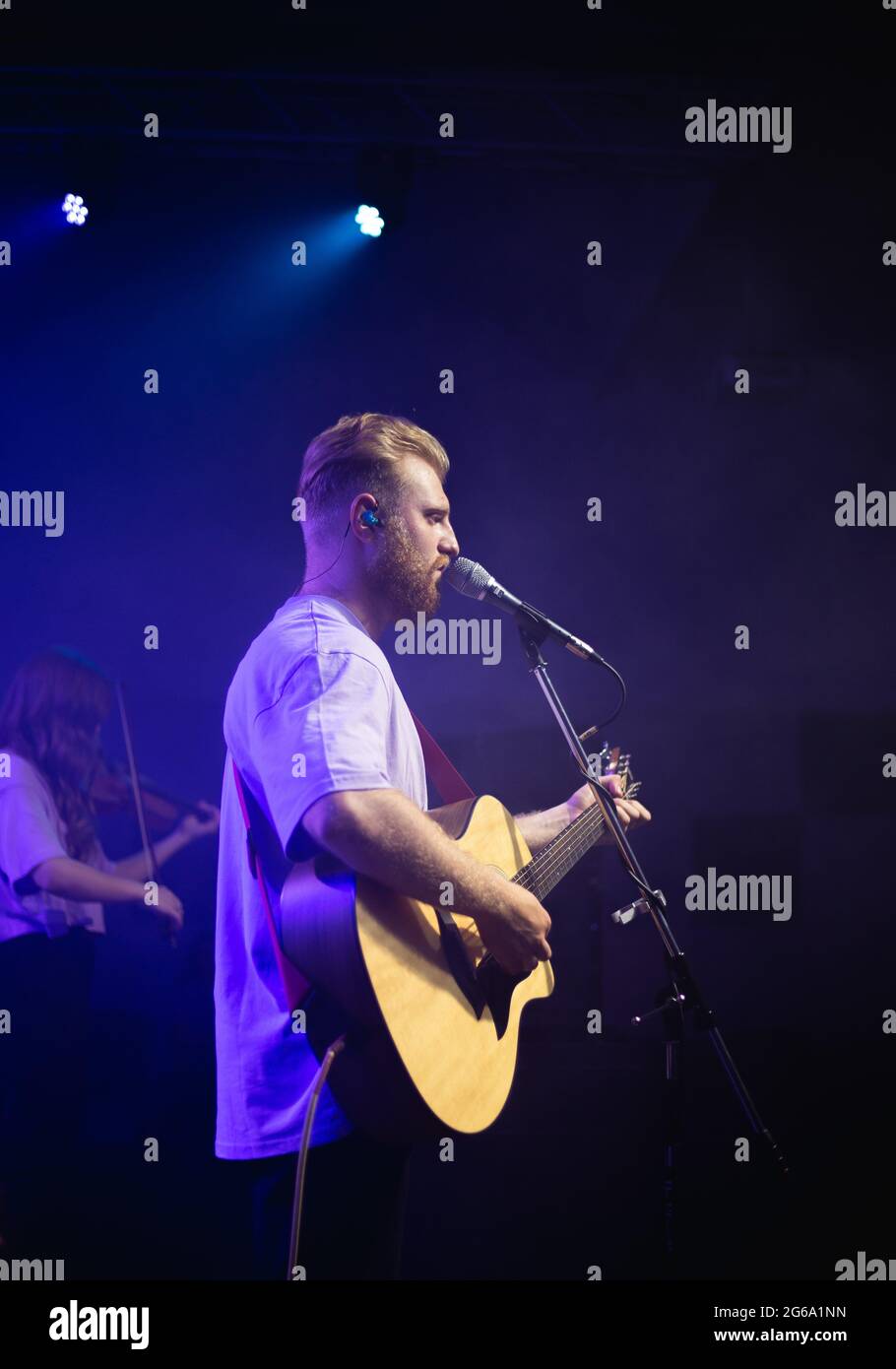 A young man in a white T-shirt with a beard holds an acoustic guitar in his hand and sings into a microphone stands on the stage Stock Photo