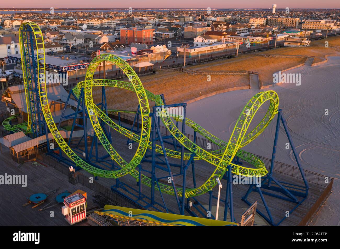Aerial of a wild, twisting, green roller coaster over the water with the beach and town in the background at sunrise. Stock Photo