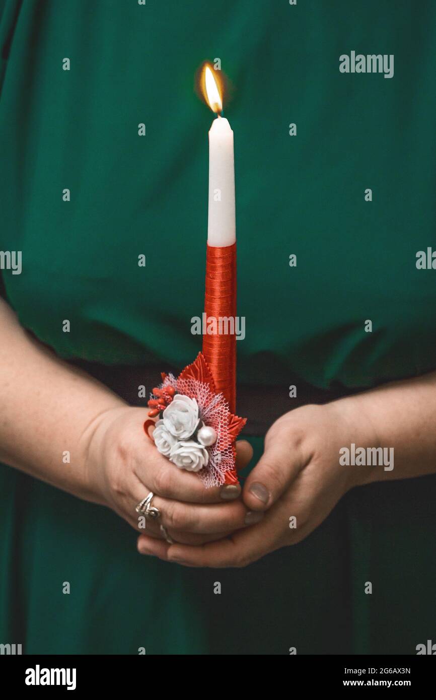 The hands of a woman in a green dress hold a large burning decorative candle, close-up. Stock Photo