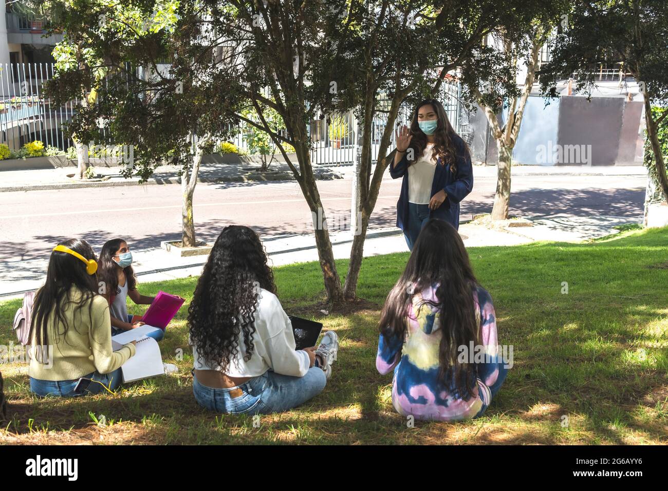 Latina teacher teaching in a face mask with her teenage students with their backs to the lesson in an outdoor classroom. Stock Photo