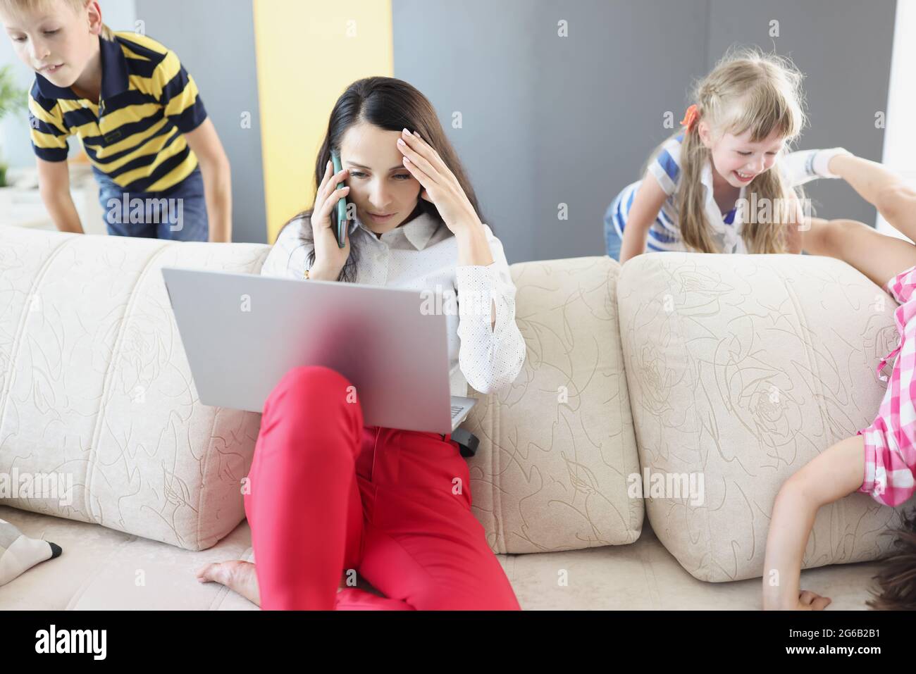 Sad young woman sitting on sofa with laptop and holding her head near children playing at home Stock Photo
