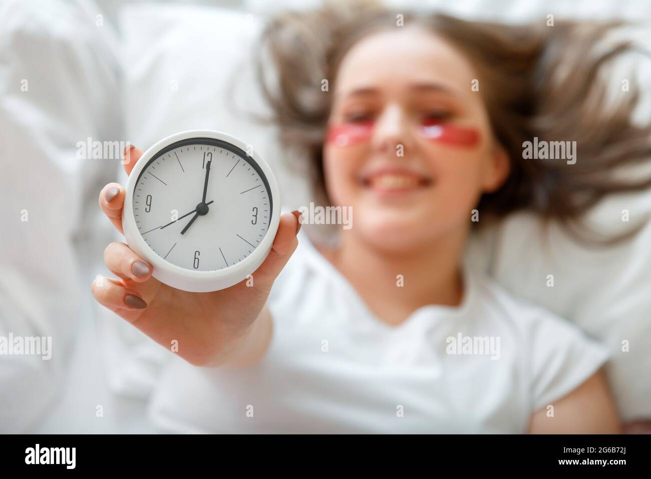 Happy young woman wake up after sleeping holding bed side alarm clock in hand. Morning beauty routine in pajamas. Woman use eye patches after wake up Stock Photo