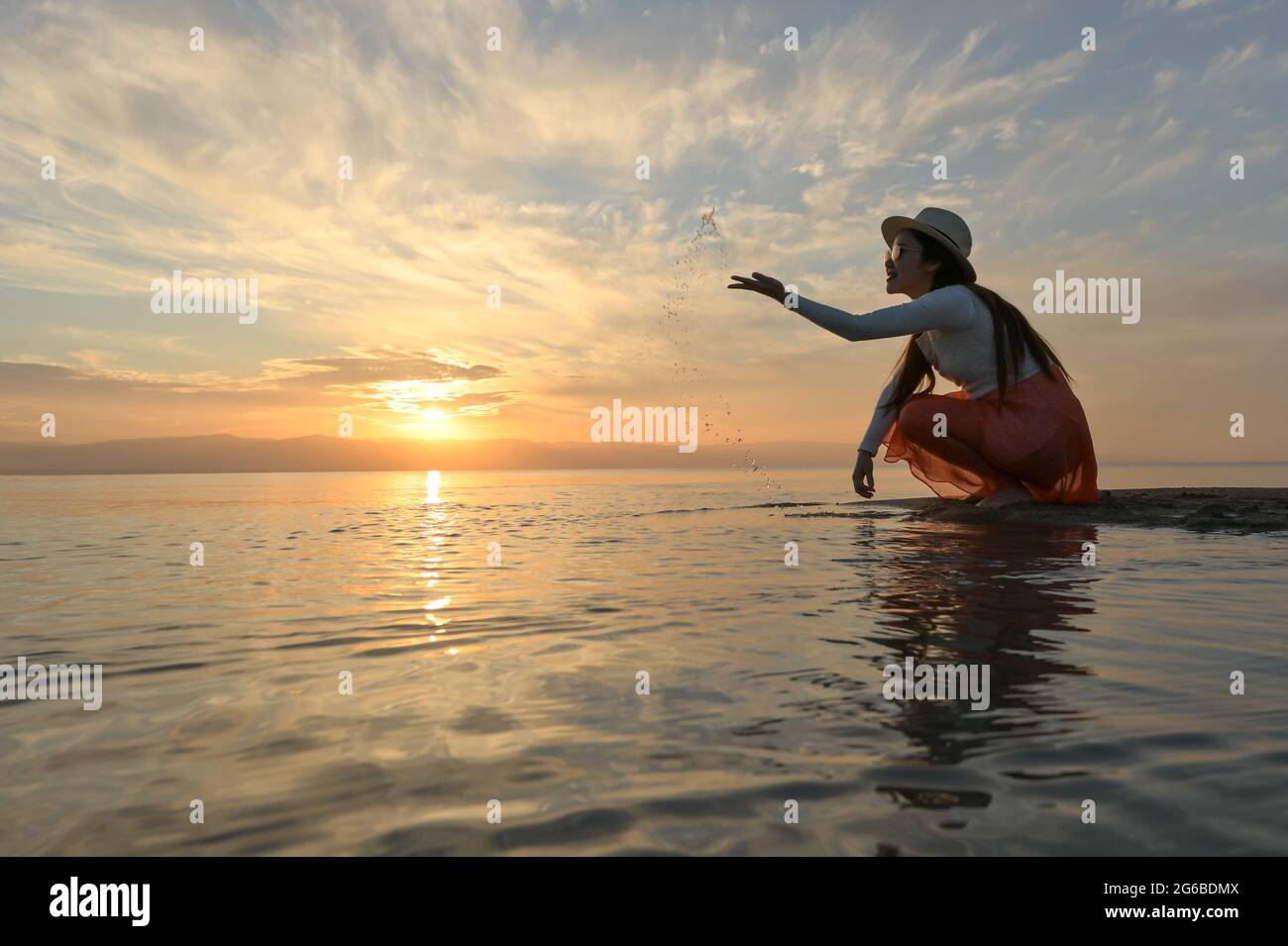 Beautiful woman crouching by sea throwing water in the air at sunset, Thailand Stock Photo