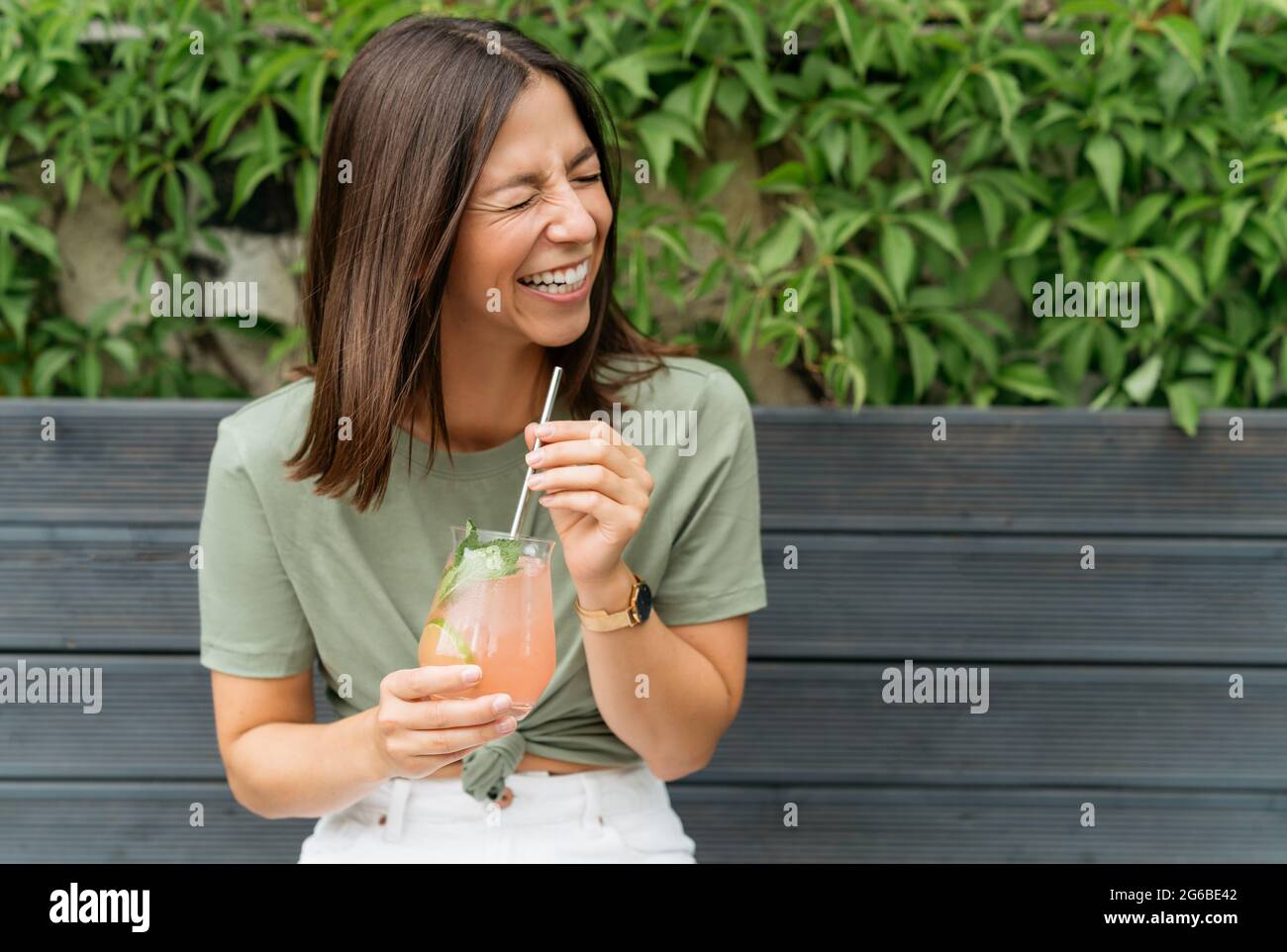 Portrait of a woman with holding a paloma cocktail sitting on a bench laughing Stock Photo