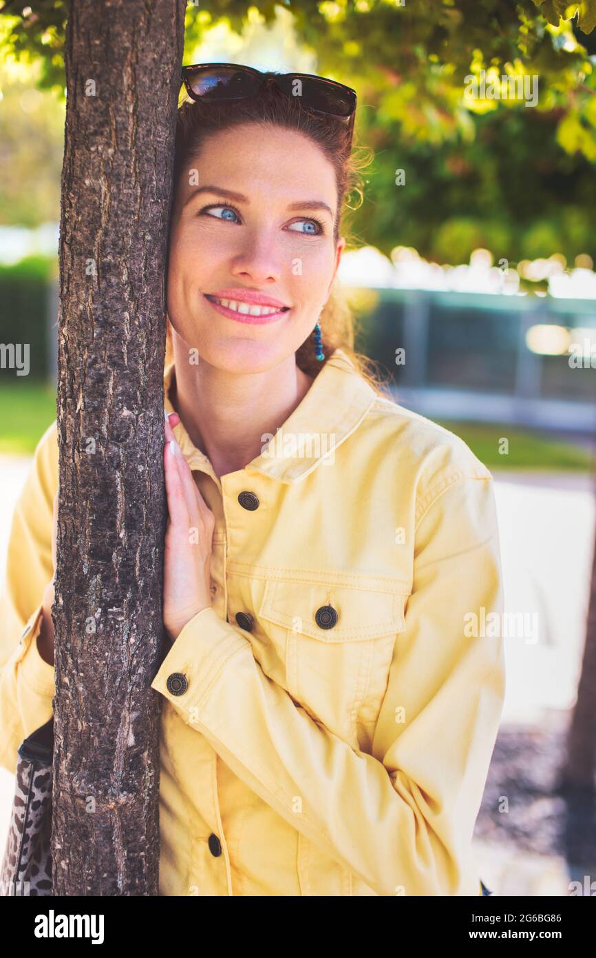 Happy young Caucasian romantic woman thinking about love in park at tree, outdoors Stock Photo