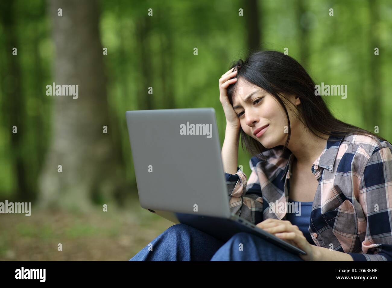 Mixed race sad woman checking laptop sitting in a forest Stock Photo