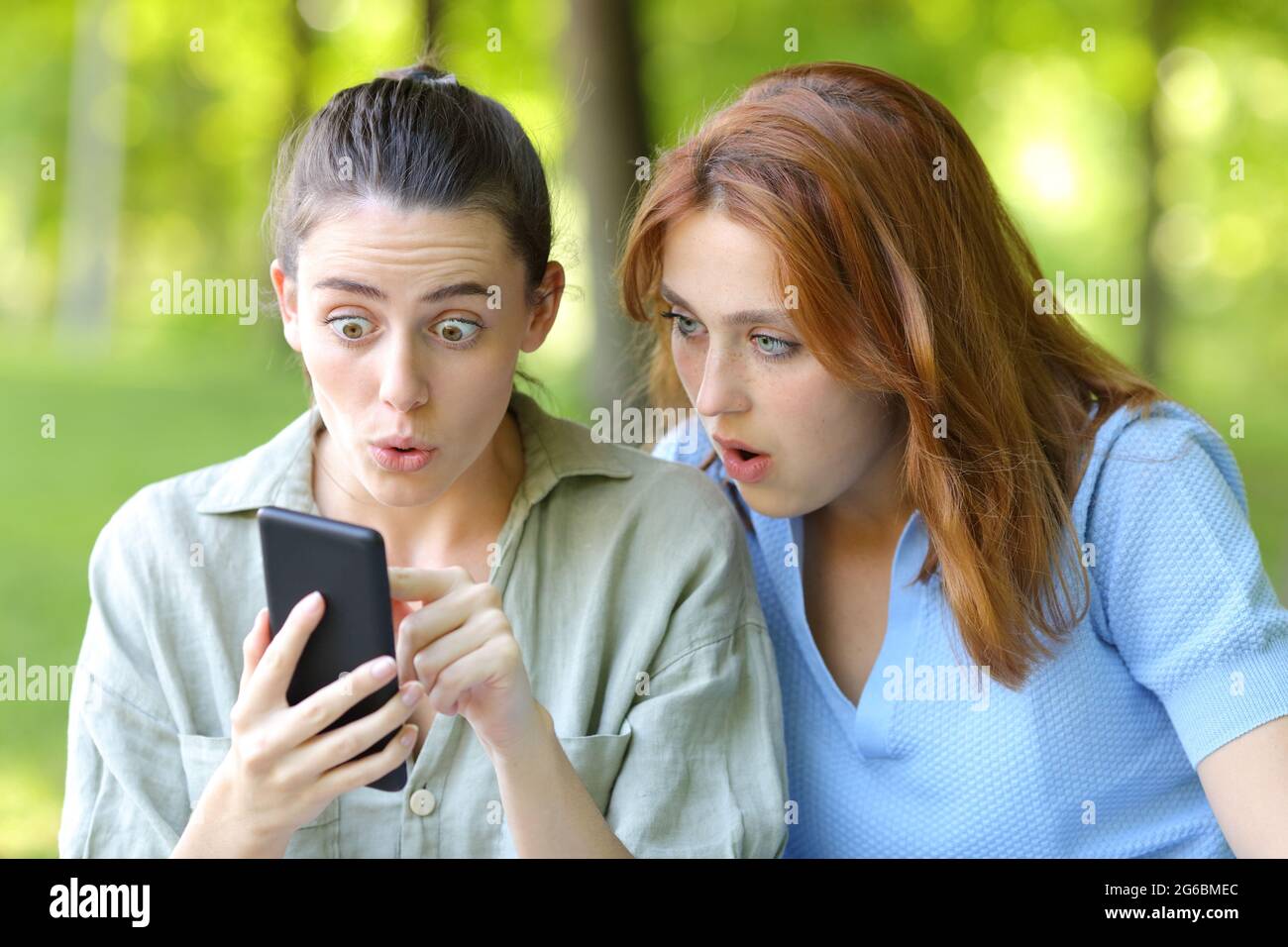 Two amazed women checking mobile phone content in a park Stock Photo