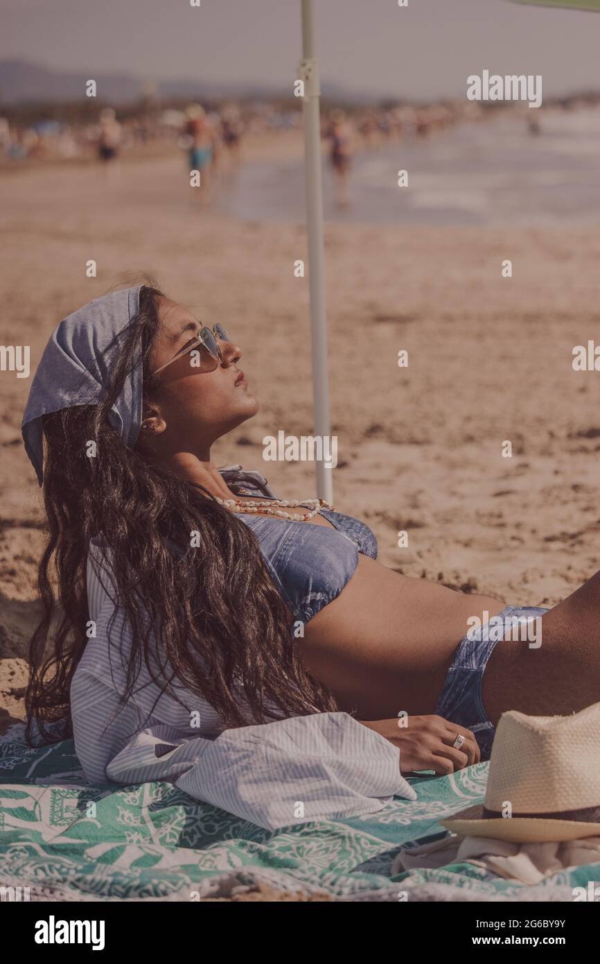 Vertical view black indian girl wearing bikini and sunglassess lying on her beach towel getting tan on the beach. Summer Vibes Concept 2021. Stock Photo