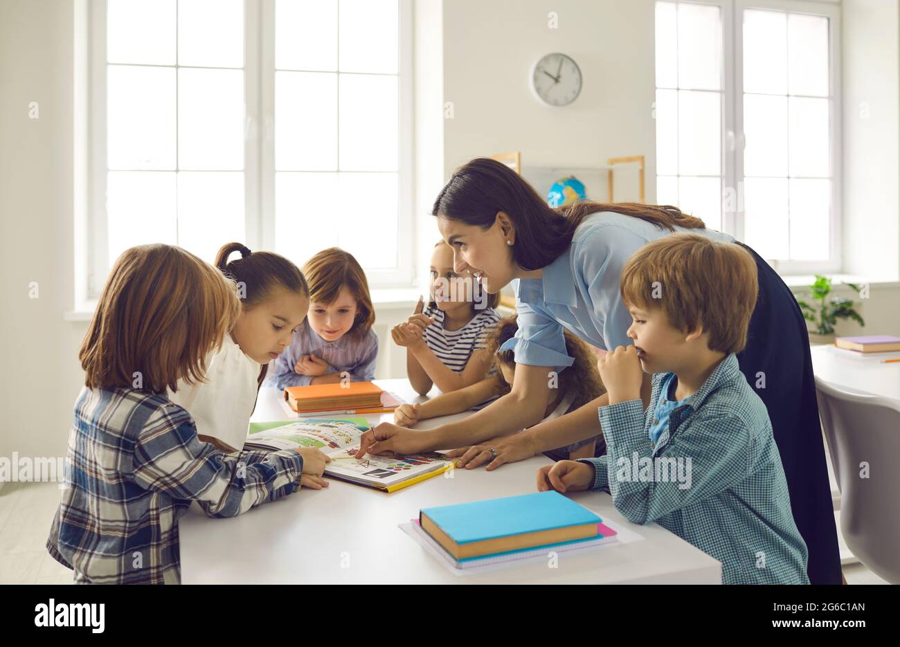 Happy school teacher and children standing around table and reading books together Stock Photo
