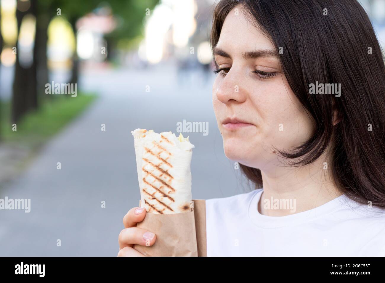 Brunette woman overeats shawarma on a city street. Street fast food pita roll with meat and vegetables. Stock Photo