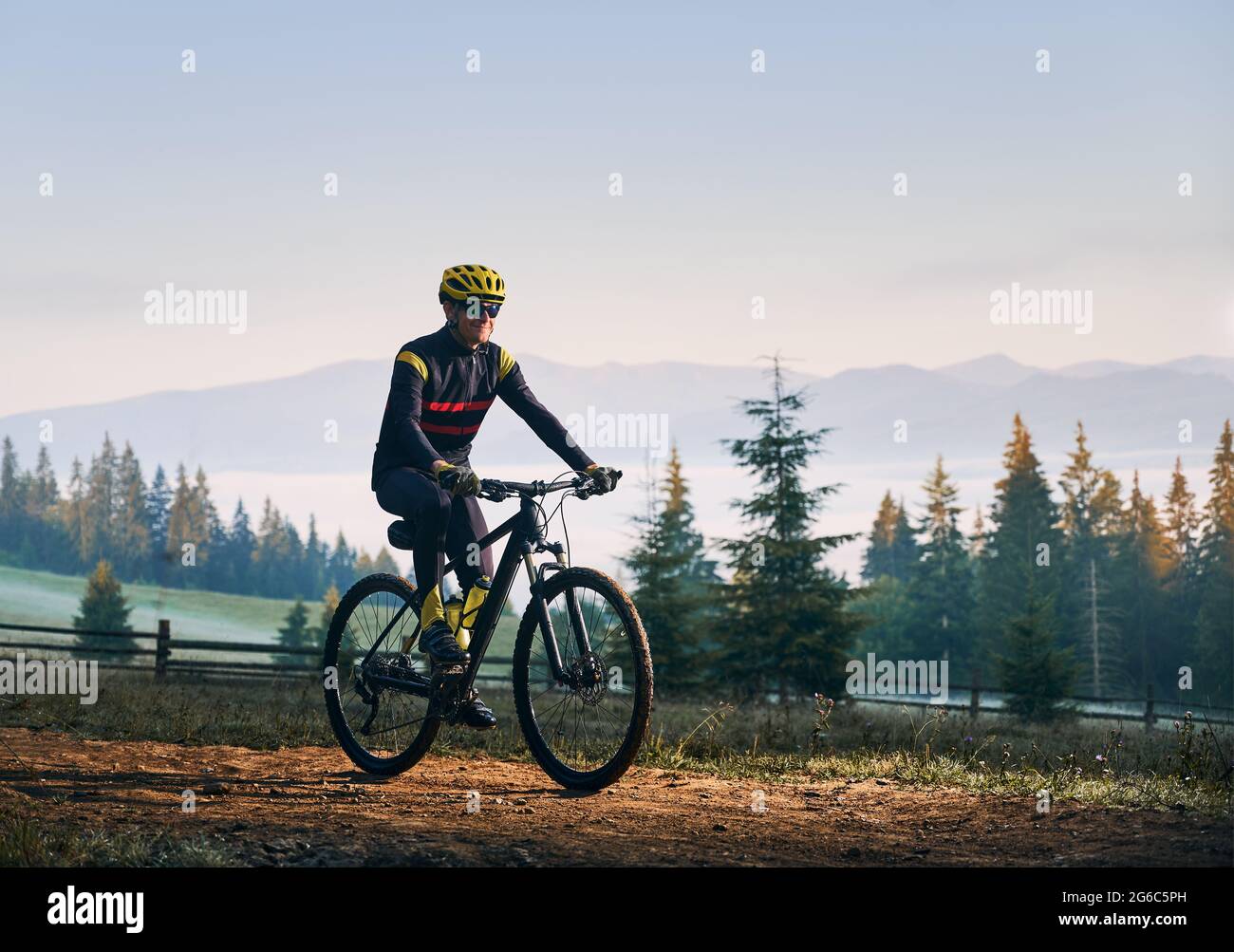 Happy cyclist in cycling suit riding bike on mountain road with coniferous trees and hills on background. Man bicyclist wearing safety helmet and glasses while enjoying bicycle ride in mountains. Stock Photo