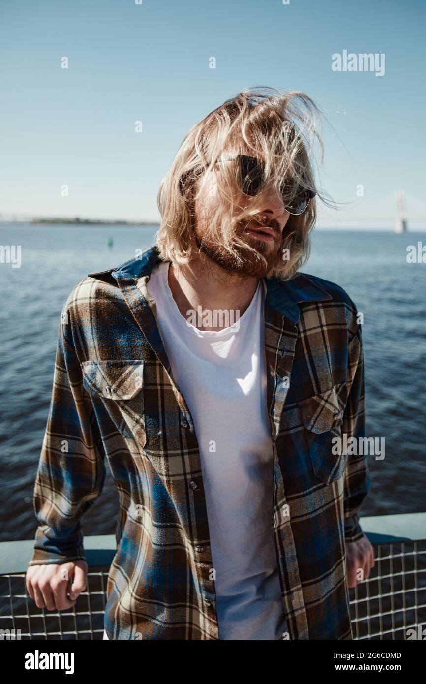 Vertical film portrait of long haired young man wearing sunglasses while posing by river in wind Stock Photo