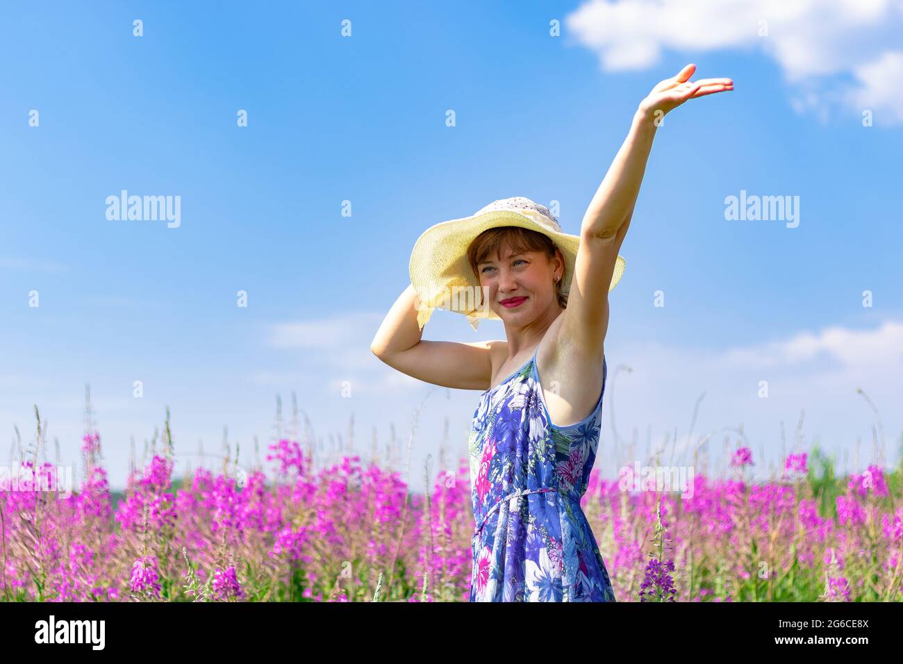 Young woman in a field of flowers in a straw hat against a background of green forest and blue sky on a hot summer day. Selective focus Stock Photo