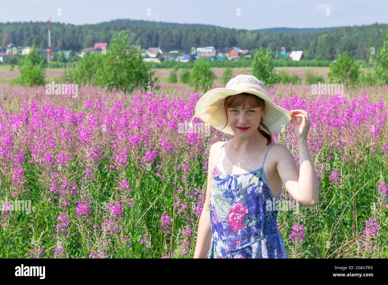 Young woman in a field of flowers in a straw hat against a background of green forest and blue sky on a hot summer day. Selective focus Stock Photo