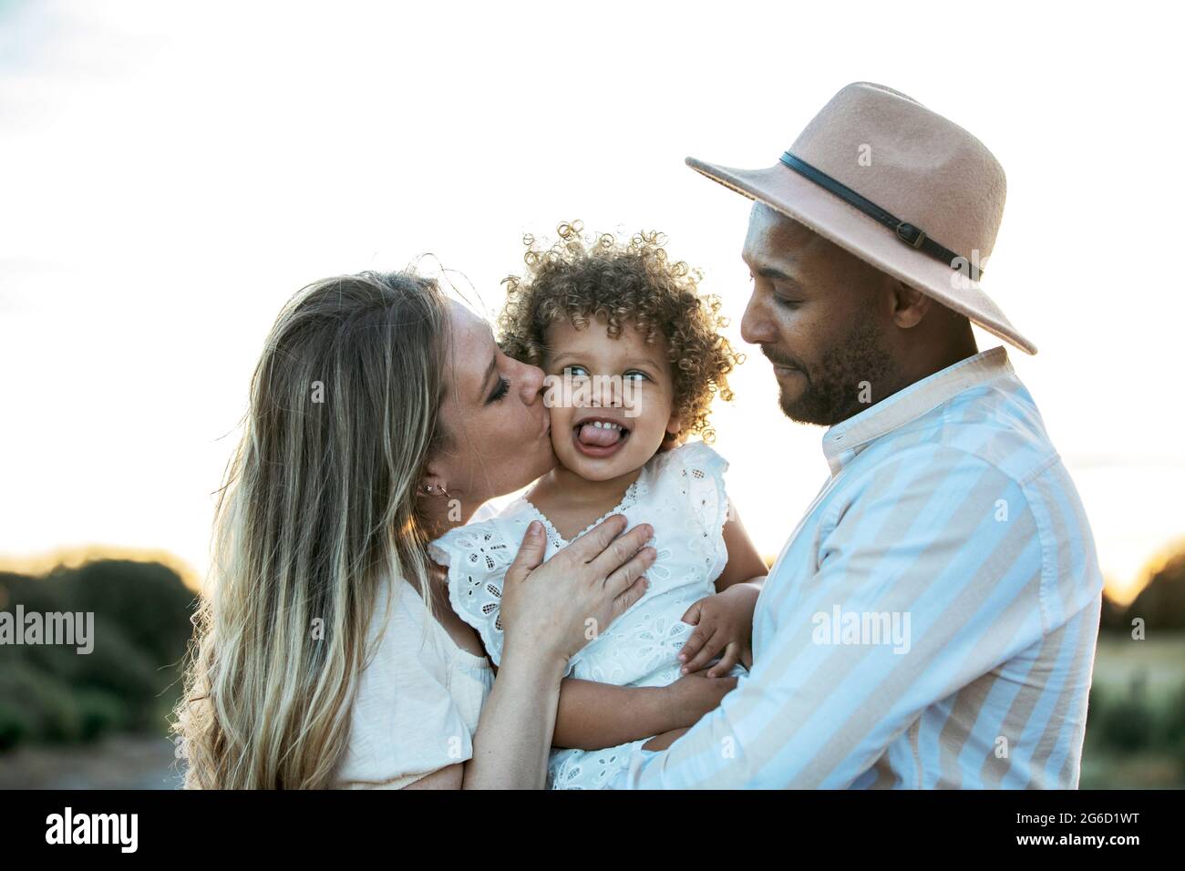 Delighted multiethnic family hugging cute little daughter while standing against sunset in field in summer Stock Photo