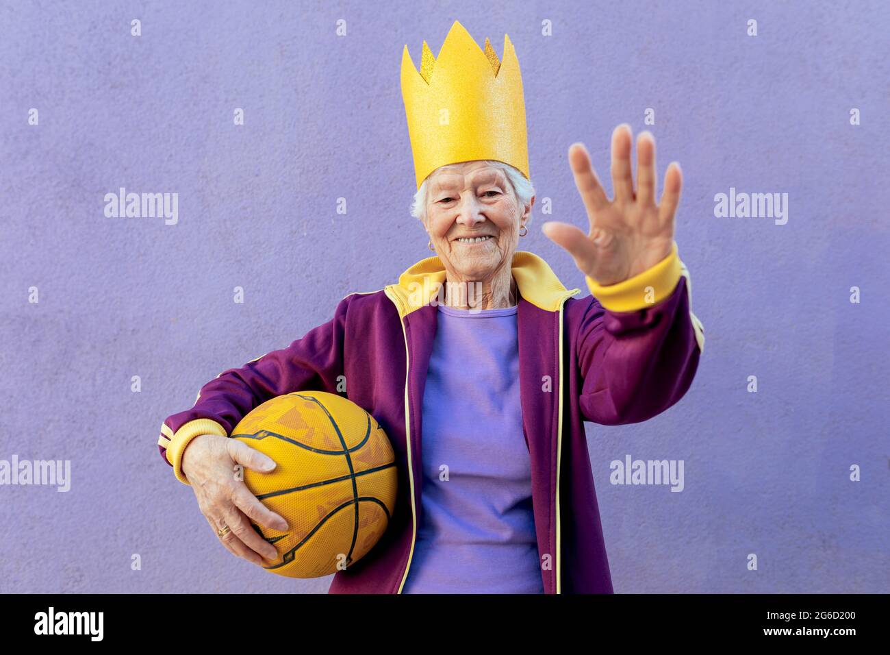 Cheerful senior female athlete in sportswear with basketball looking at camera while demonstrating control gesture on purple background Stock Photo