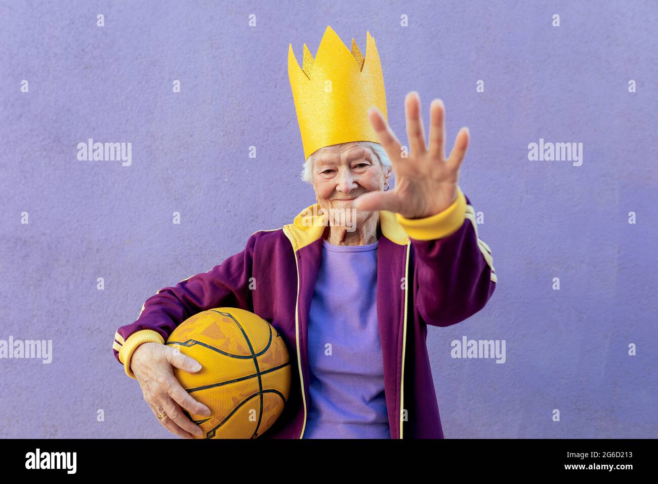 Cheerful senior female athlete in sportswear with basketball looking at camera while demonstrating control gesture on purple background Stock Photo