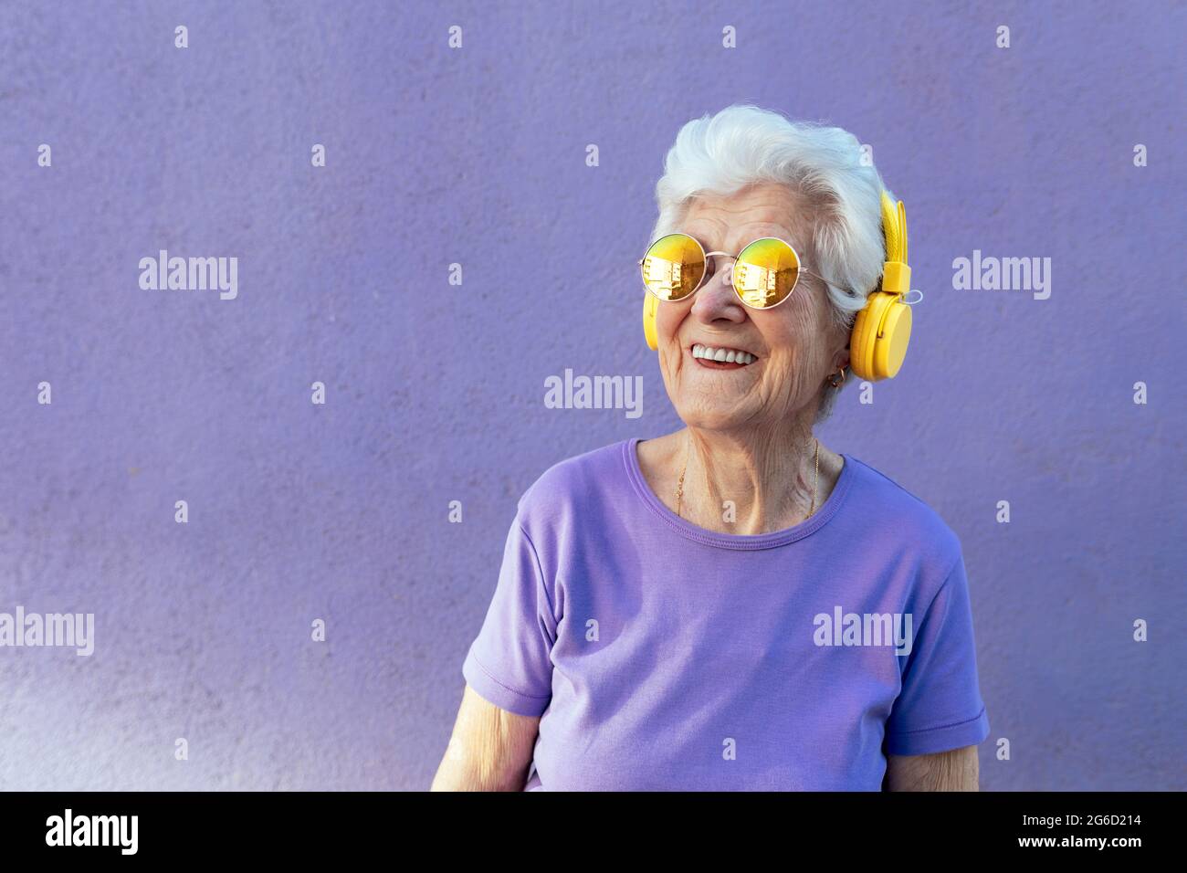 Cheerful senior female in t shirt and modern sunglasses listening to song from wireless headset on purple background Stock Photo