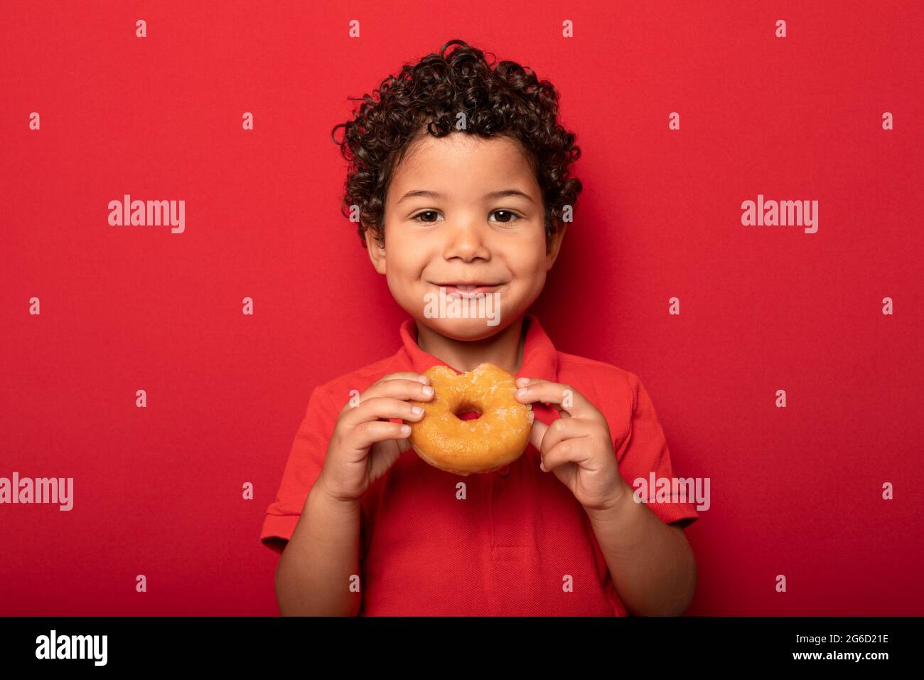 Adorable child with curly hair eating sweet tasty doughnut and looking at camera on red background Stock Photo