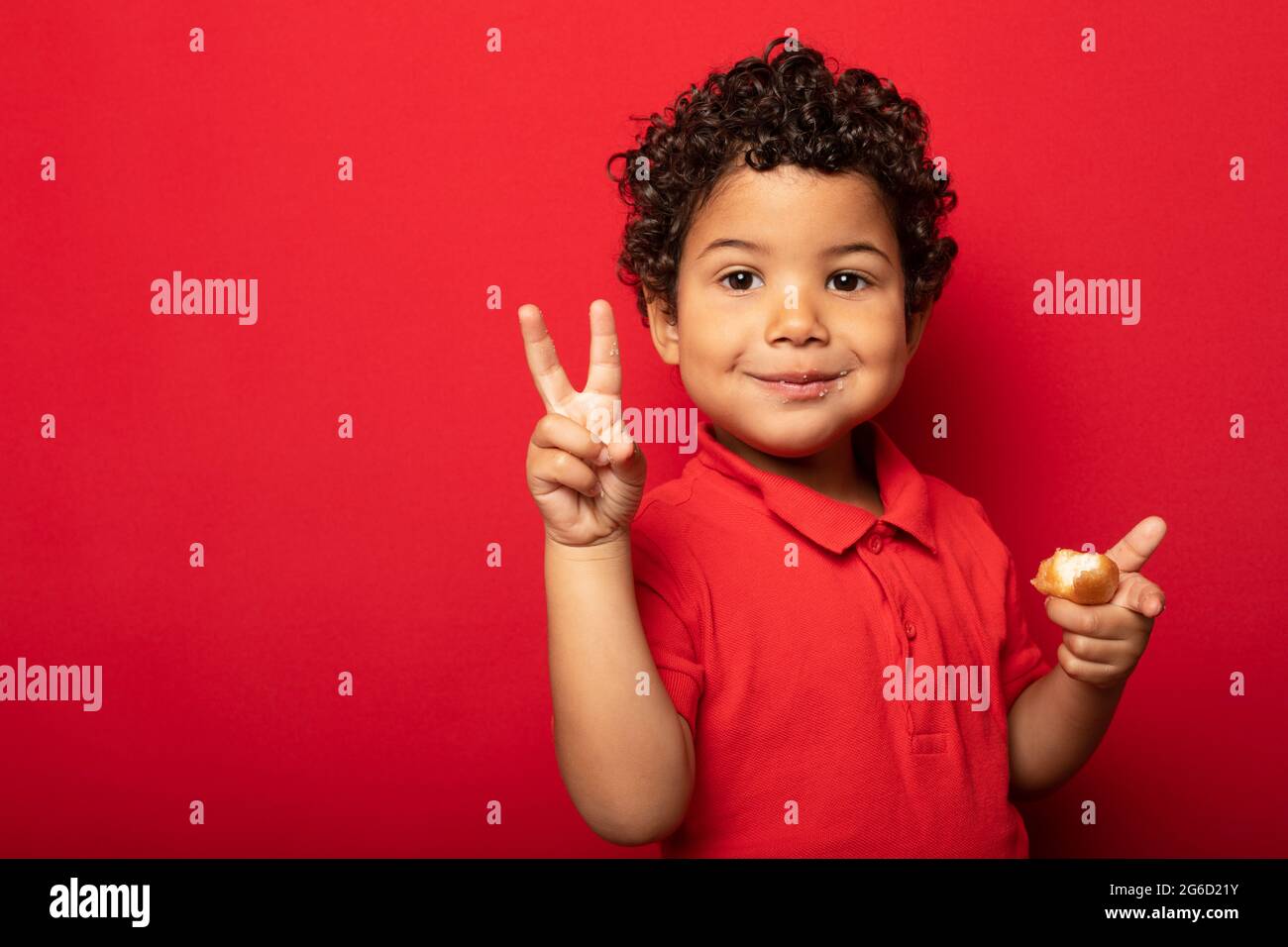 Adorable child eating delicious doughnut and showing V sign while looking at camera on red background in studio Stock Photo
