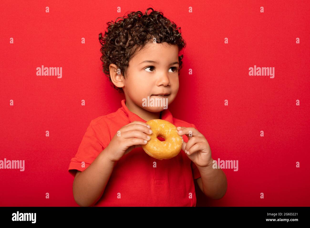 Adorable child with curly hair eating sweet tasty doughnut and looking away on red background Stock Photo