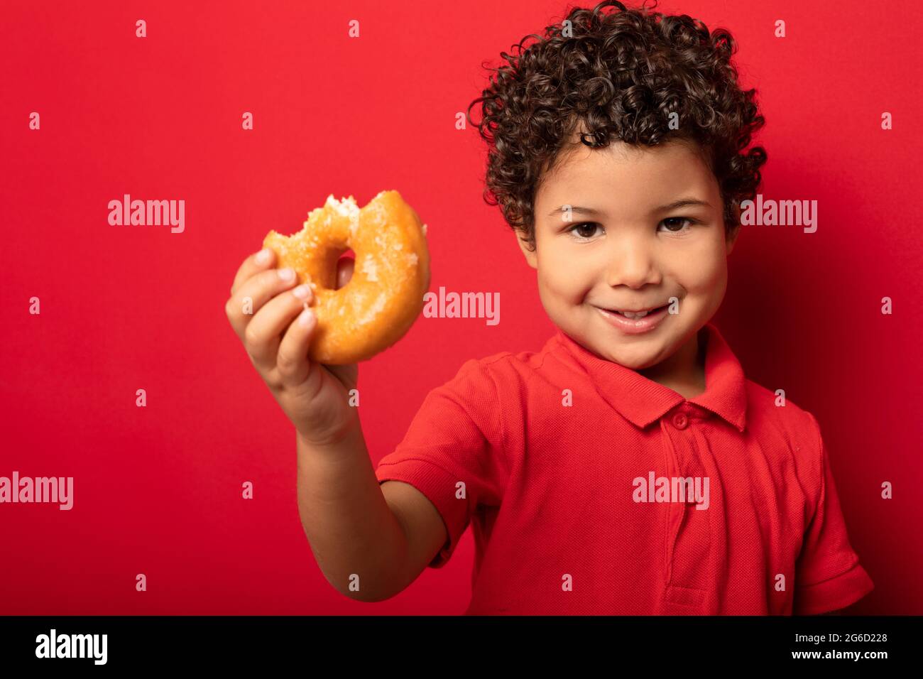 Adorable child with curly hair eating sweet tasty doughnut and looking at camera on red background Stock Photo