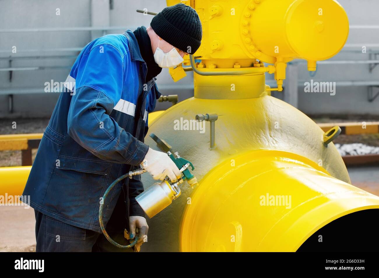 A painter in working clothes paints a metal shut-off valve for gasification from a compressor gun on a summer day. Professional painting of parts. Industrial background. Stock Photo