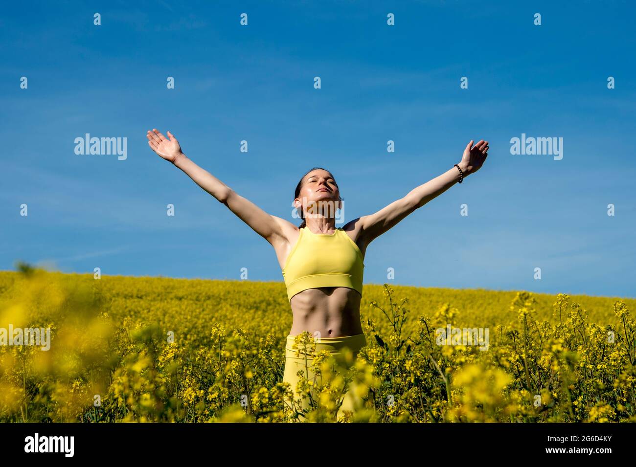 fit sporty woman wearing yellow activewear standing in a yellow fied with her arms raised enjoying the sun Stock Photo