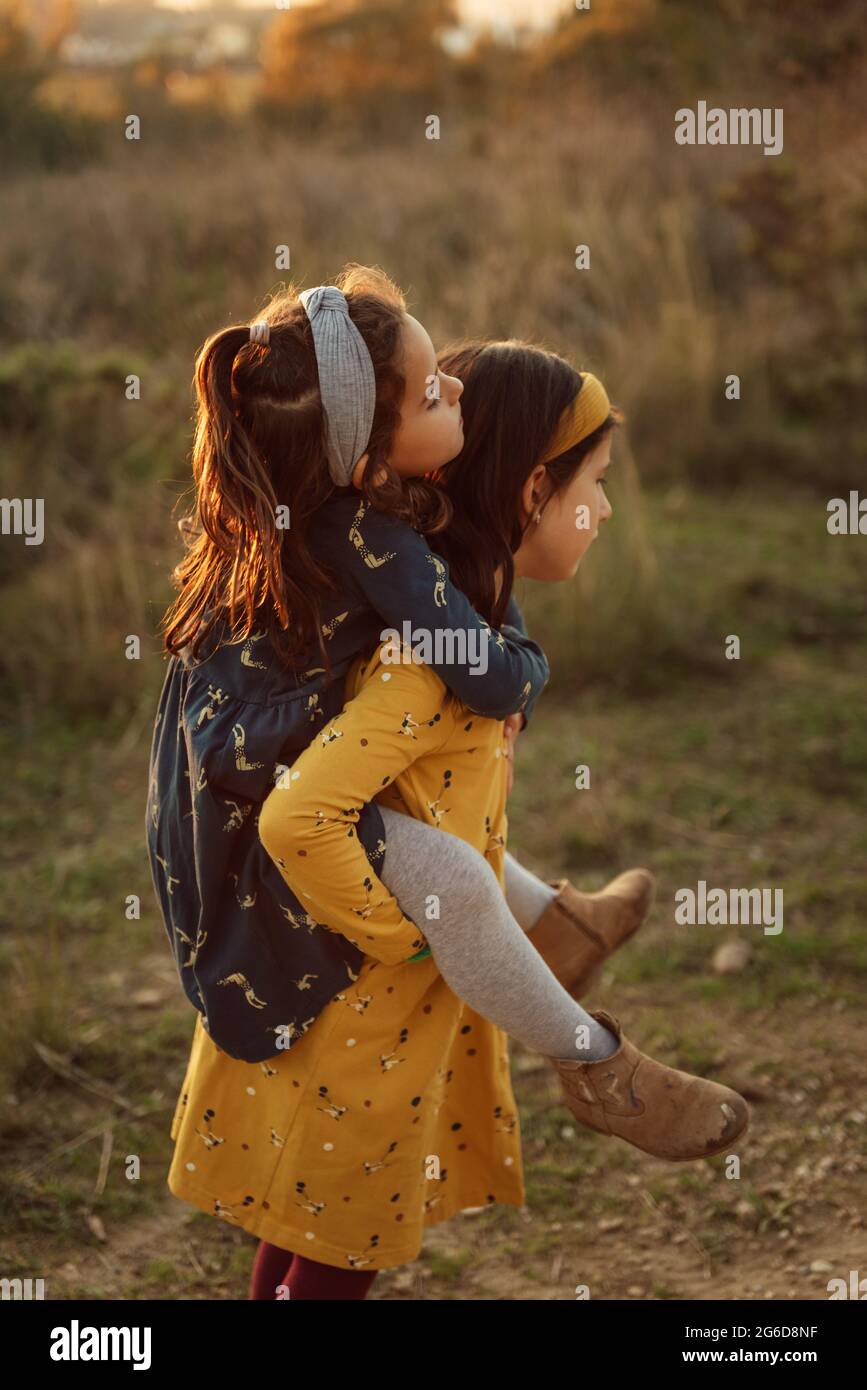 Side view of loving preteen girl carrying cute little sister on back while spending summer day together in field Stock Photo