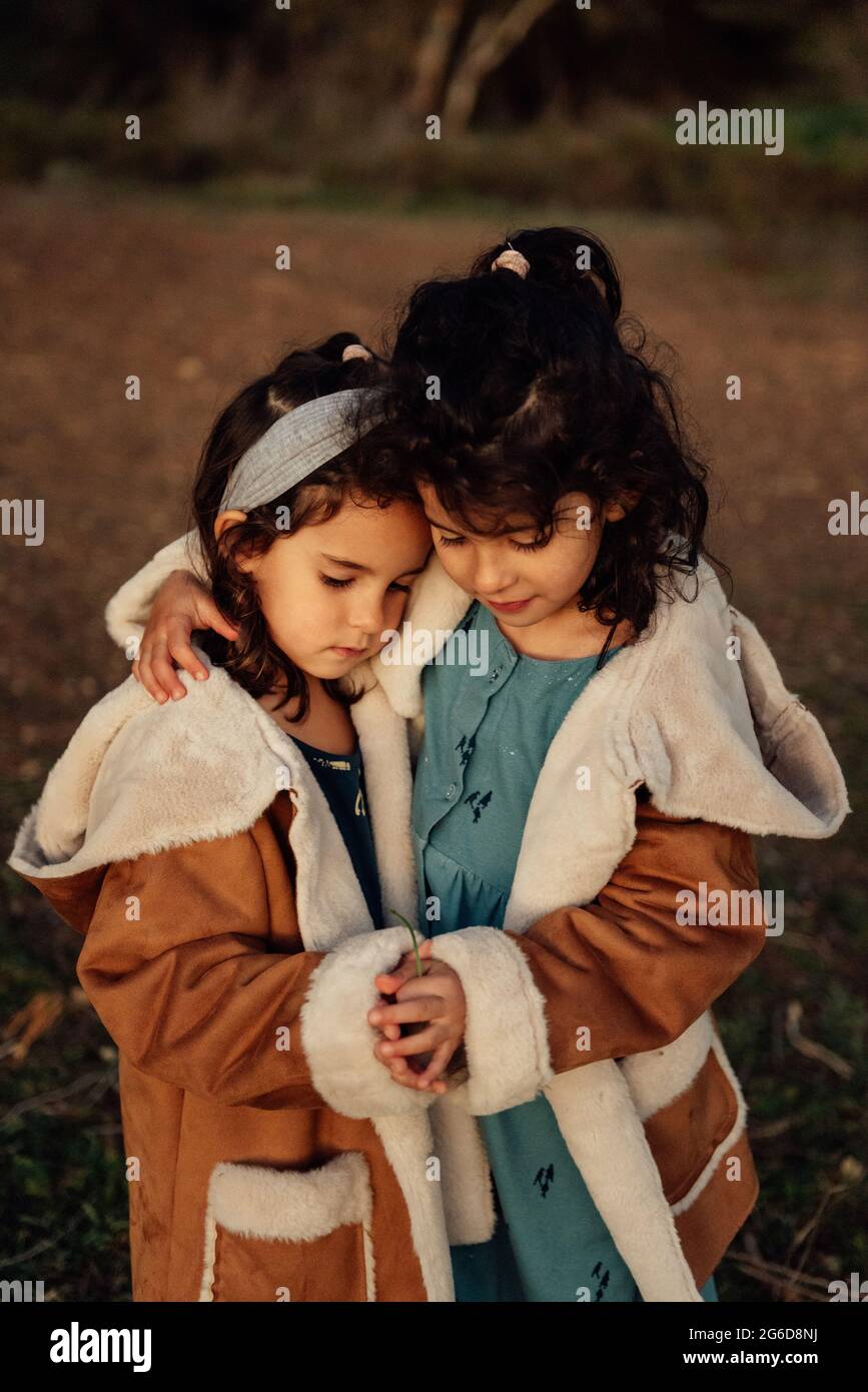 Cute peaceful little sisters in similar outerwear embracing each other while standing together against blurred background of nature Stock Photo