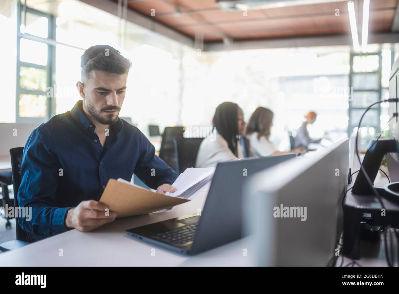 Concentrated male employee sitting at table with laptop and reading documents while working in spacious office with blurred colleagues Stock Photo