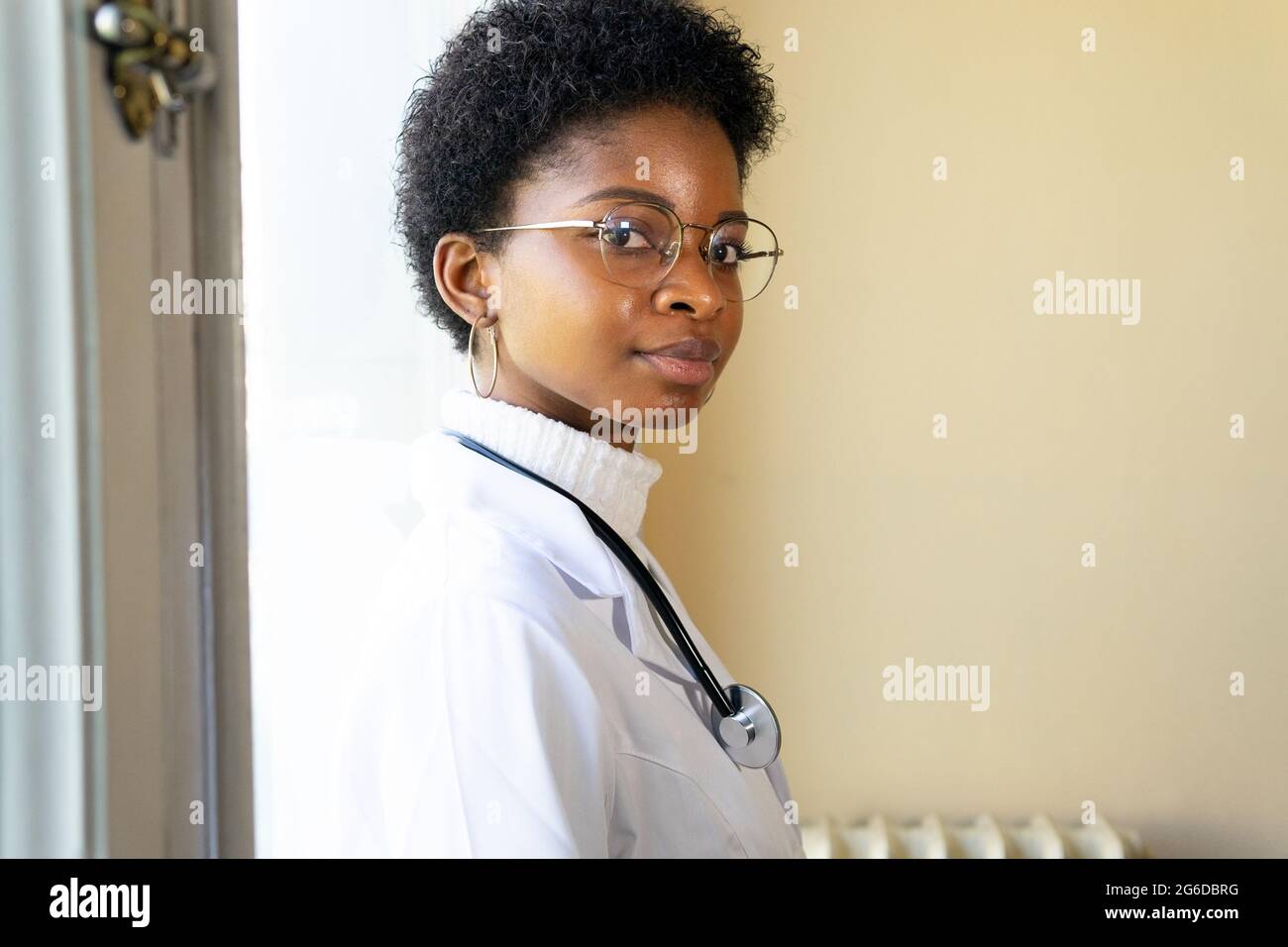 Side view of competent young African American female physician in white medical coat and eyeglasses with stethoscope looking at camera while standing Stock Photo