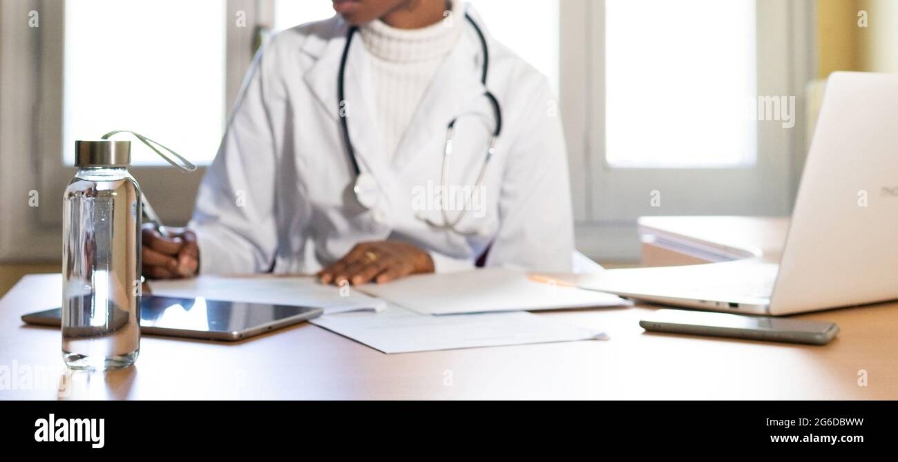 Crop unrecognizable black female physician with stethoscope writing information on paper sheet while preparing medical report at table in office of mo Stock Photo