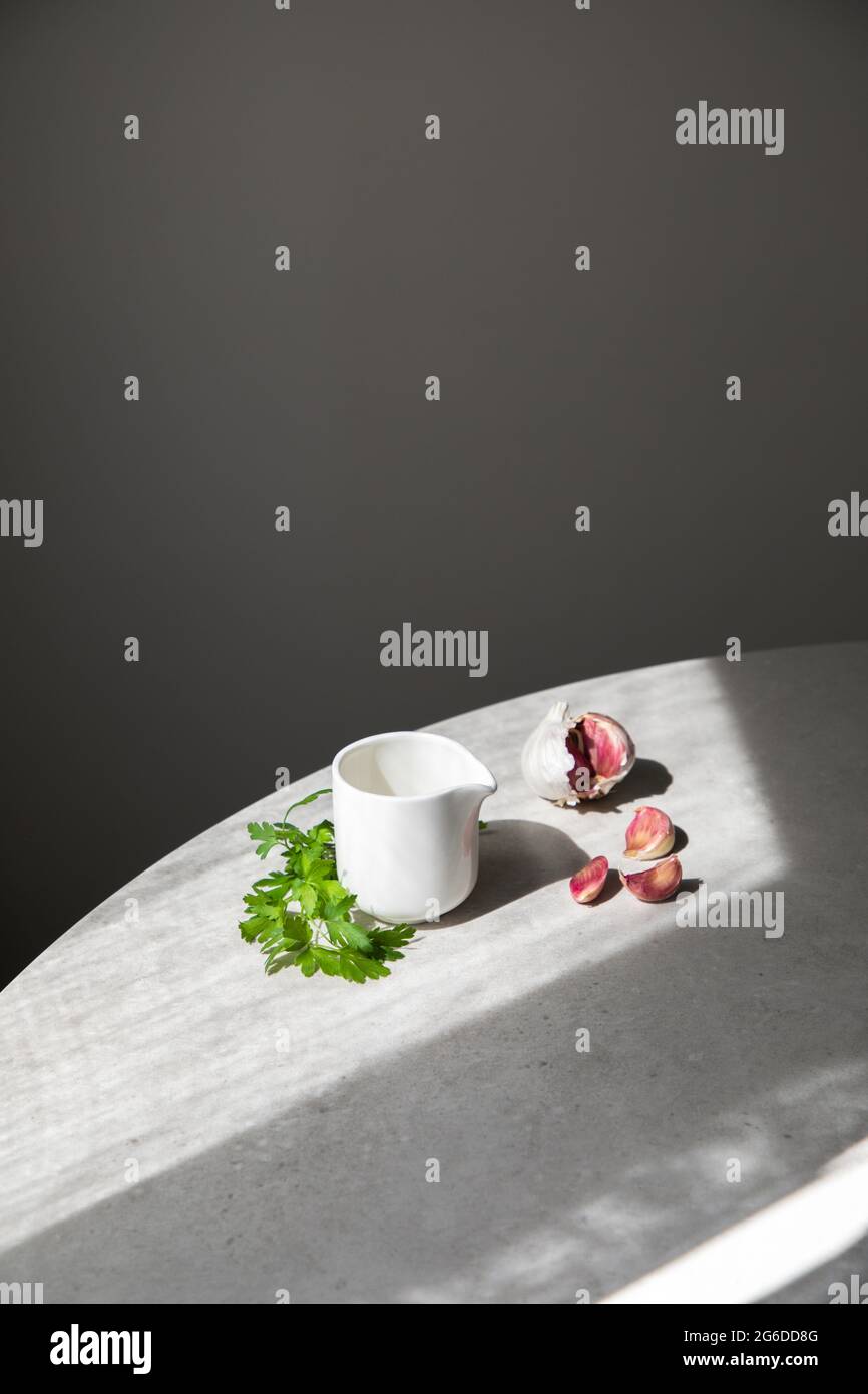 From above garlic cloves and fresh green parsley placed on round table near ceramic sauce boat on gray background Stock Photo