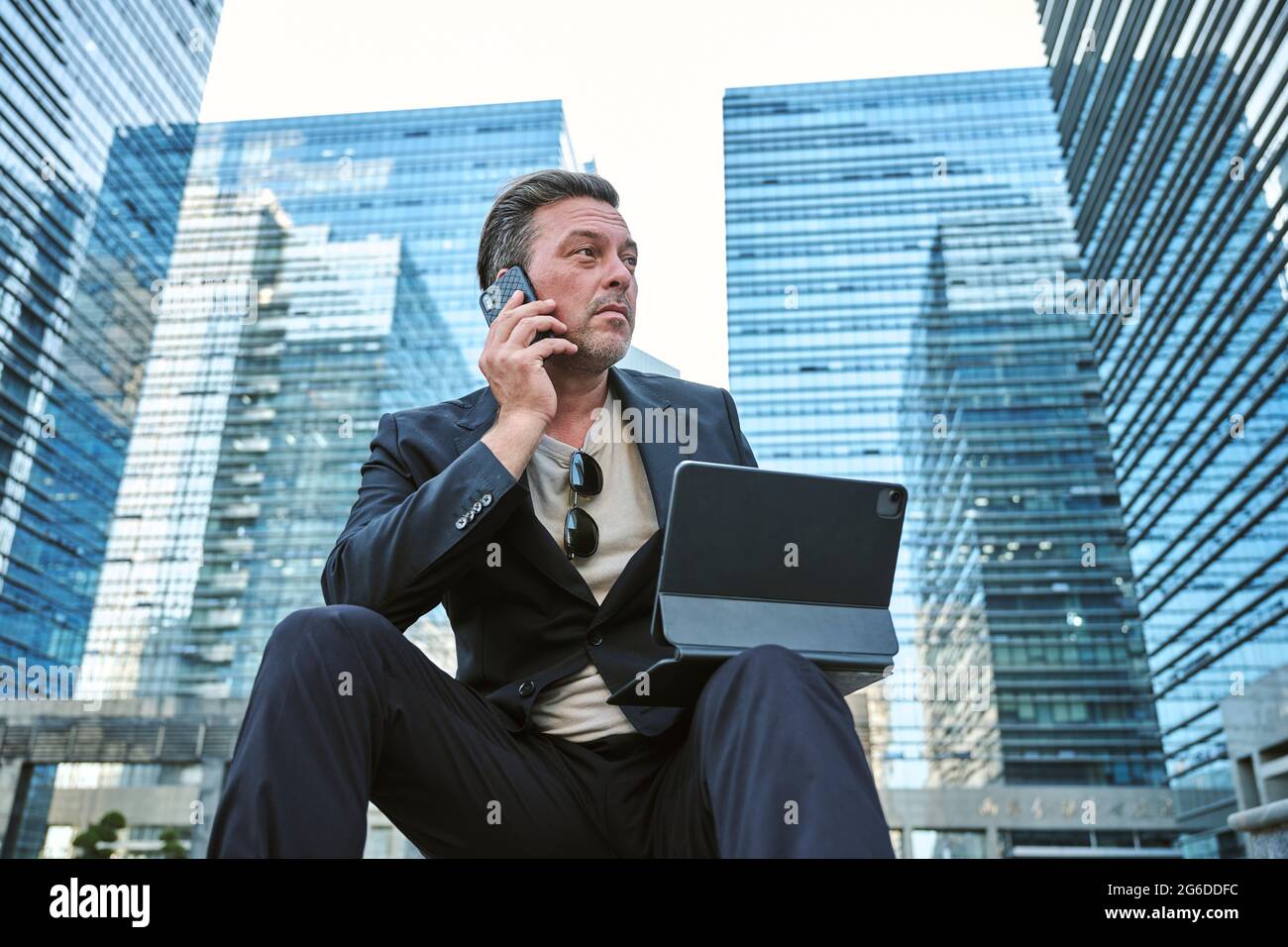 Business man having a call on his smartphone and working with his tablet while sitting around office's buildings Stock Photo