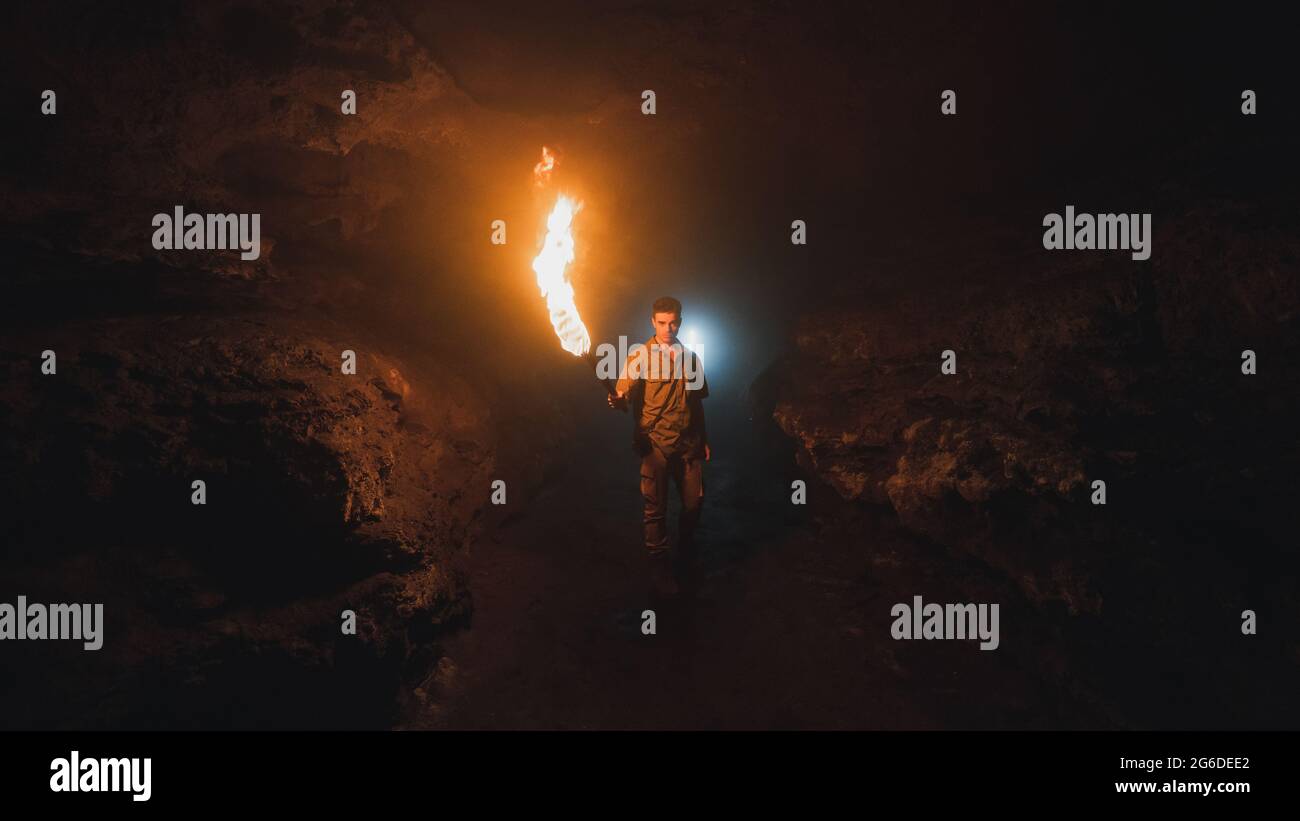 Young male speleologist with flaming torch standing in dark narrow rocky cave while exploring subterranean environment looking at camera Stock Photo