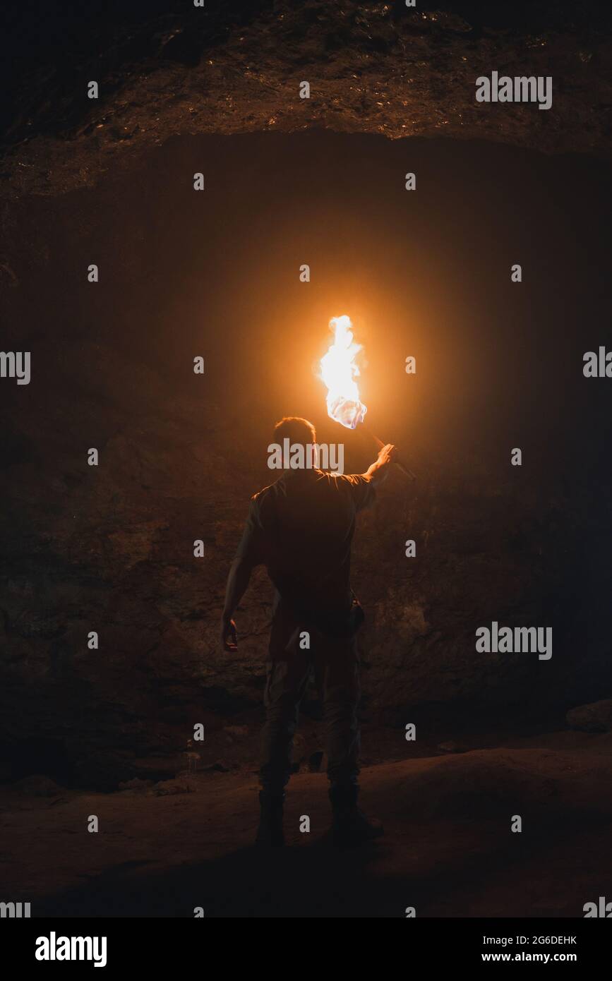 Back view of unrecognizable male speleologist with flaming torch standing in dark narrow rocky cave while exploring subterranean environment Stock Photo