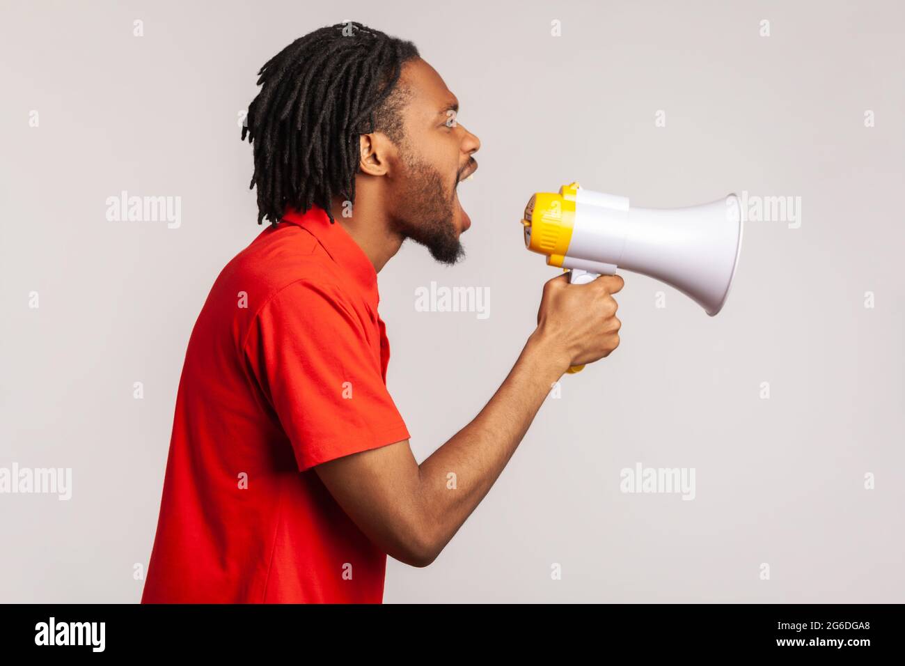 Profile of man with dreadlocks and beard wearing red casual style T-shirt, loudly screaming at megaphone, making protest, wants to be heard. Indoor st Stock Photo