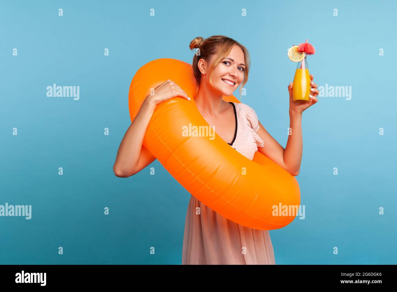 Happy blonde woman with orange rubber ring rising hand with cocktail and looking directly at camera with charming smile, wearing summer dress. Indoor Stock Photo
