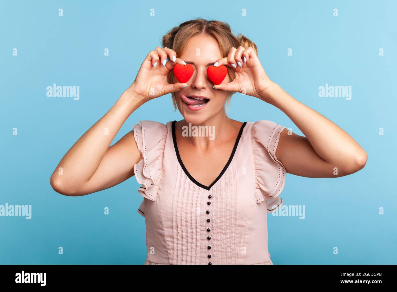 Loving eyes. Beautiful young adult woman with two funny hair buns holding two valentine hearts in front of her eyes like glasses, shows tongue out. In Stock Photo